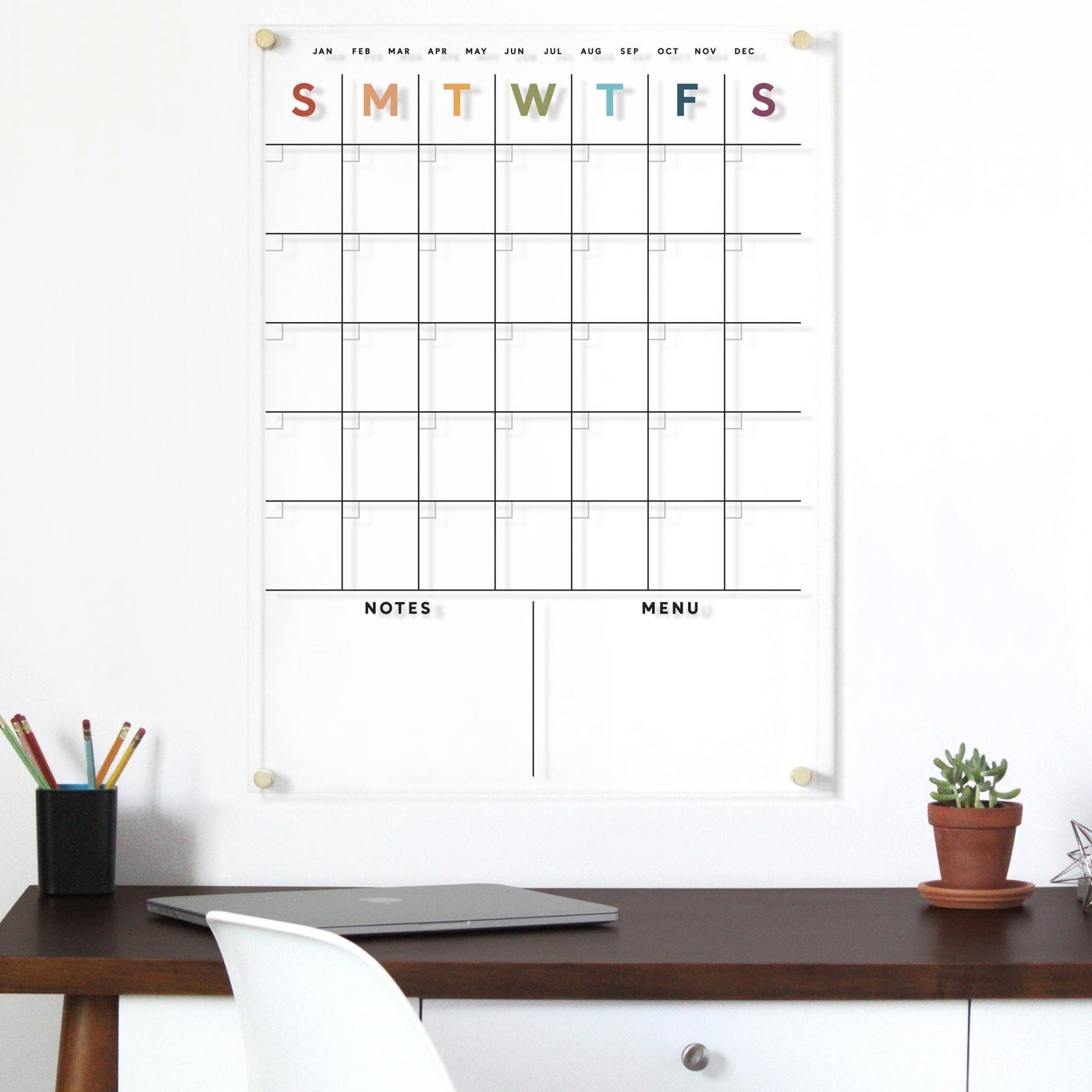 The Clear Acrylic Monthly Wall Calendar by Girl Friday features days in earth tone rainbow letters and two sections for notes and menu planning. A dark wooden desk holds a closed laptop, pencil holder with colored pencils, and a white chair. Enjoy high-quality materials for lasting style.
