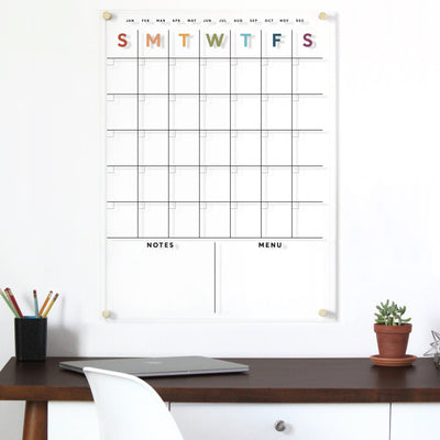 The Clear Acrylic Monthly Wall Calendar by Girl Friday features days in earth tone rainbow letters and two sections for notes and menu planning. A dark wooden desk holds a closed laptop, pencil holder with colored pencils, and a white chair. Enjoy high-quality materials for lasting style.