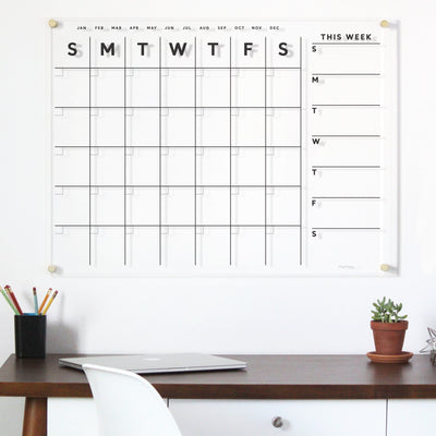 A Clear Acrylic Customizable Monthly & Weekly Wall Calendar from Girl Friday adorns the wall, complemented by a desk with a white chair, pencils in a cup, and a small potted plant. This chic setup is ideal for planning with wet-erase markers, blending style and function seamlessly.