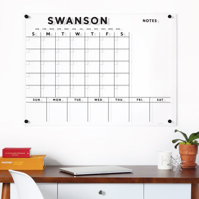A Wall-mounted Clear Acrylic Customizable Monthly & Weekly Wall Calendar by Girl Friday with SWANSON at the top displays days and months. Below, a desk holds colorful books, a silver laptop, a white mug with wet-erase markers, and a potted plant.