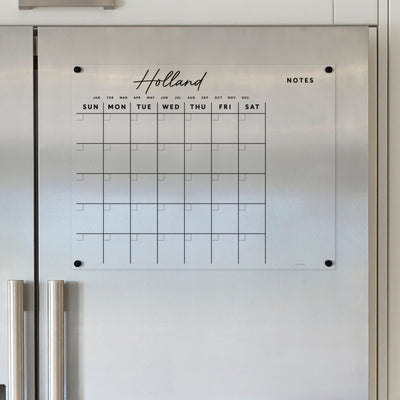 The elegant Acrylic Fridge Calendar with Custom Family Name | Horizontal by Girl Friday features an acrylic wall mount, English days/months, weekly rows, and note space. Ideal above kitchen appliances with a white backdrop, its perfect for wet-erase markers.
