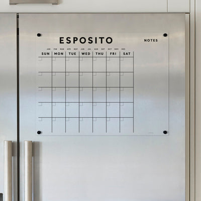 The Girl Friday Acrylic Fridge Calendar with Custom Family Name in black text is mounted on a gray metallic surface. It features a weekly grid labeled Sunday to Saturday and includes space for notes, designed for use with dry-erase markers.