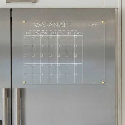 The Girl Friday Acrylic Fridge Calendar, customizable with your family name like Watanabe and featuring white text, includes a grid for days, months, and a Notes section. It comes with wet-erase markers and complements sleek stainless steel kitchen appliances when mounted on a gray wall.