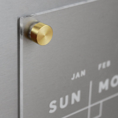 The Girl Friday Clear Acrylic Monthly Fridge Calendar features a modern design with clear acrylic, brass hardware, and a silver background. It displays days like Sun, Mon, and months such as Jan, Feb in white sans-serif font, ideal for wet-erase markers. Customizable with your family name!.