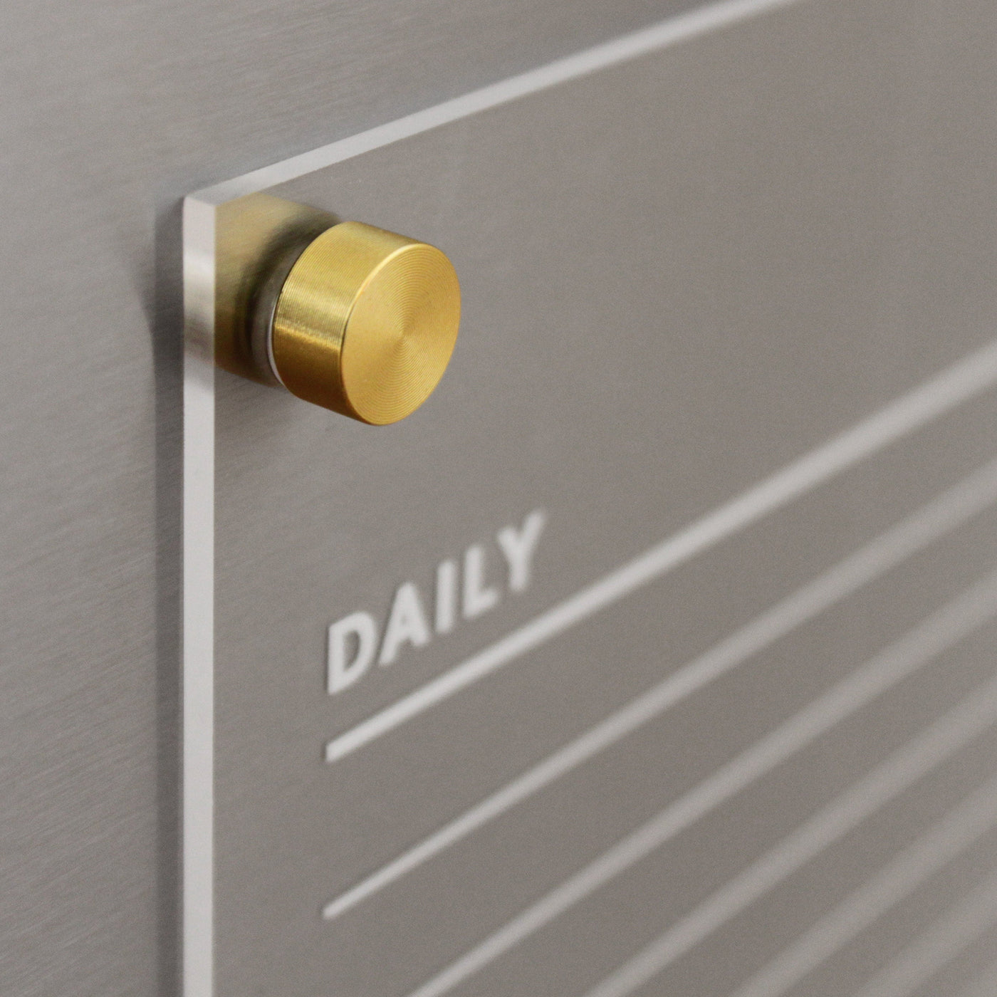 Close-up of a Girl Friday Acrylic Fridge Chore Chart for 2 children with DAILY in white. The board, ideal for wet-erase markers, features horizontal lines and is mounted with a round, golden knob against a metallic surface.