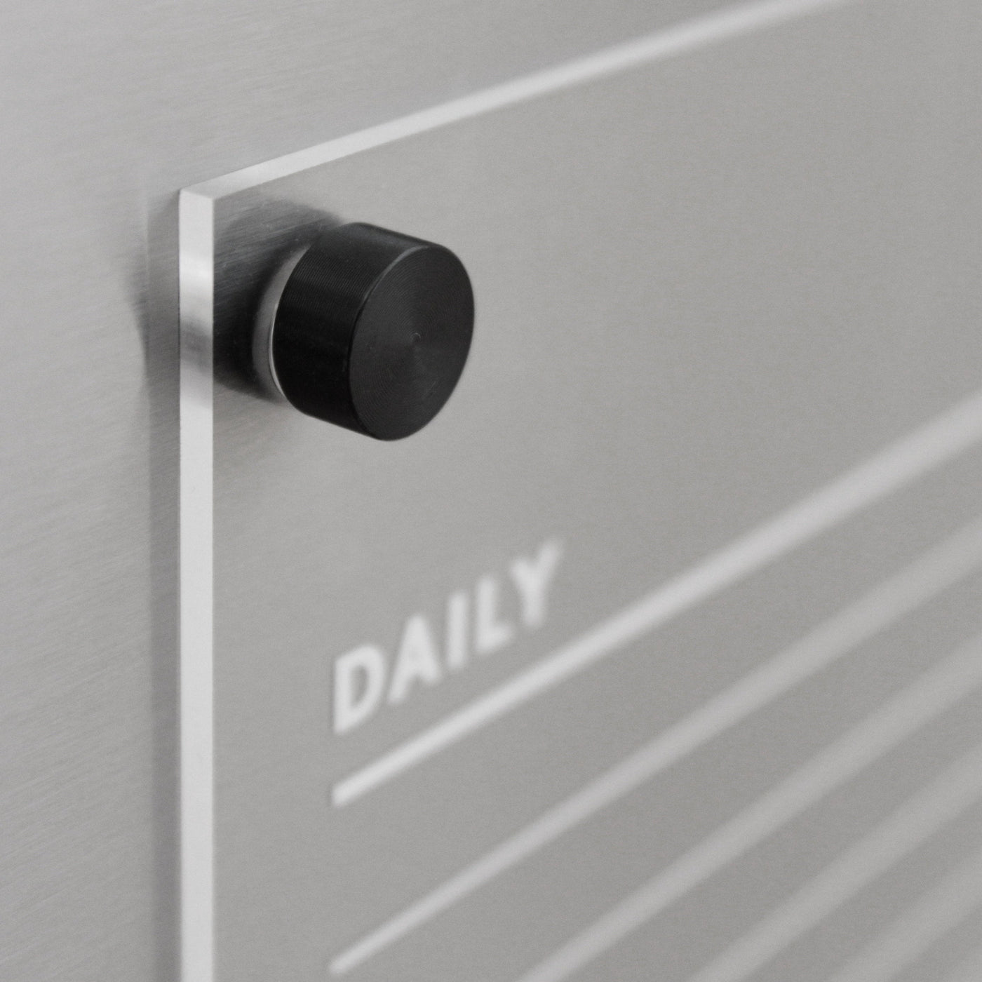 Close-up of a sleek wall organizer featuring a black knob. The word DAILY appears on a clear panel resembling the Girl Friday Acrylic Fridge Chore Chart for 2 children with white text. Horizontal lines suggest planning space, ideal for wet-erase markers on an elegant metallic gray background.