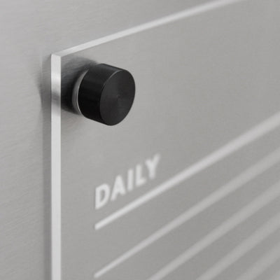 Close-up of a sleek wall organizer featuring a black knob. The word DAILY appears on a clear panel resembling the Girl Friday Acrylic Fridge Chore Chart for 2 children with white text. Horizontal lines suggest planning space, ideal for wet-erase markers on an elegant metallic gray background.