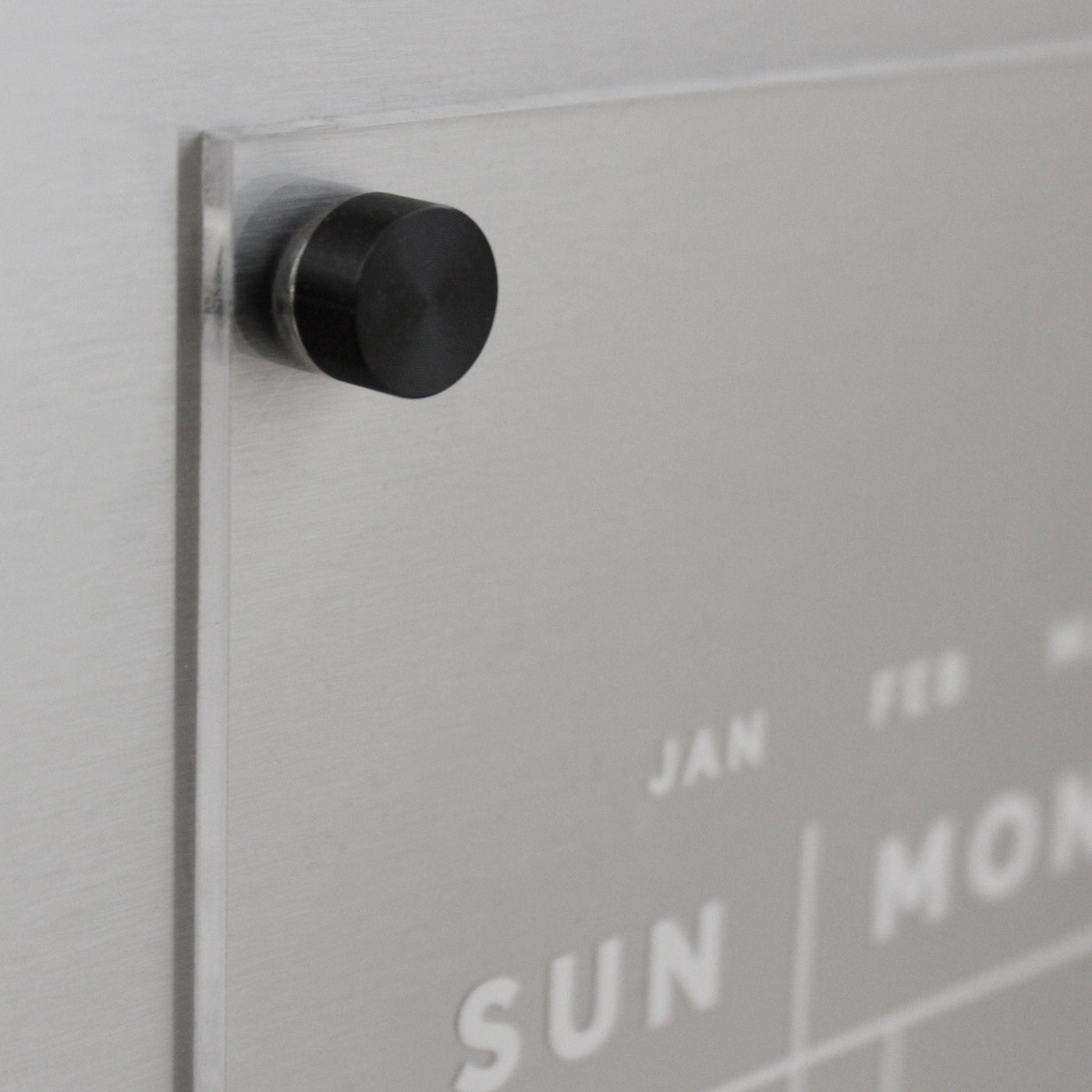 A close-up of the Girl Friday Acrylic Fridge Calendar | Custom Family Name | White Text shows a wall-mounted design with clear silver accents. The week starts on Sunday, and a black magnet holds a transparent sheet to the metal backing for easy note-taking with dry-erase markers.