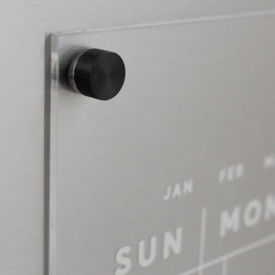 A close-up of the Girl Friday Acrylic Fridge Calendar | Custom Family Name | White Text shows a wall-mounted design with clear silver accents. The week starts on Sunday, and a black magnet holds a transparent sheet to the metal backing for easy note-taking with dry-erase markers.