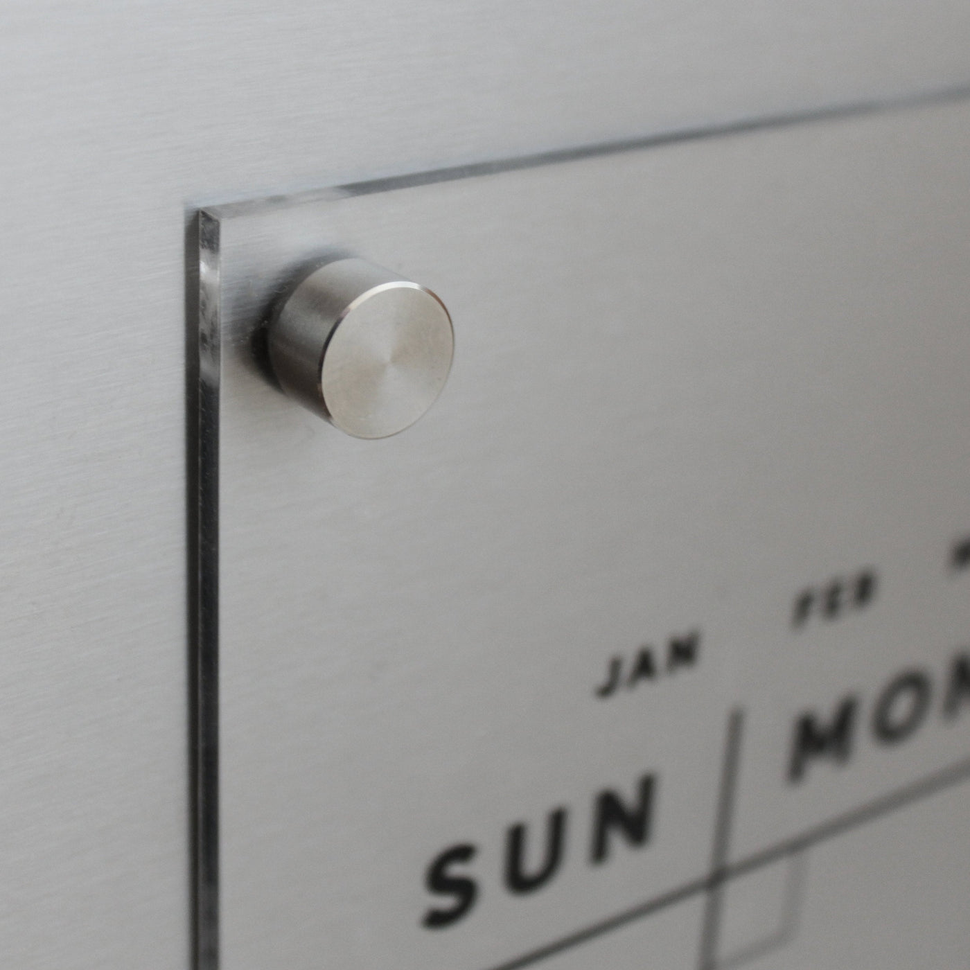 The Girl Friday Acrylic Fridge Calendar features a minimalist, transparent design with your custom family name in black text, mounted on a brushed metal background. Ideal for use with wet-erase markers, this calendar complements any modern space.