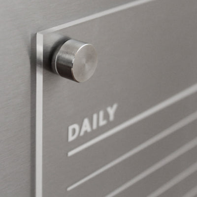 A close-up of the Girl Friday Acrylic Fridge Chore Chart for 2 children reveals a sleek board with an attached knob. It features DAILY in white text and lines for wet-erase markers. The brushed metal finish enhances its contemporary design.