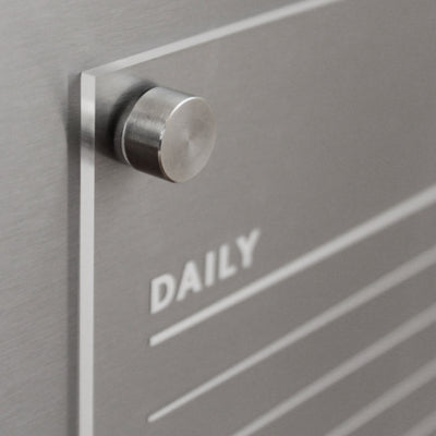 Close-up of a custom Girl Friday Acrylic Fridge Chore Chart with a metallic board, engraved lines, and the word DAILY. A metal peg casts a shadow on its minimalist, brushed silver finish. Designed for wet-erase markers. Ideal for two children with weekly notes in block font.