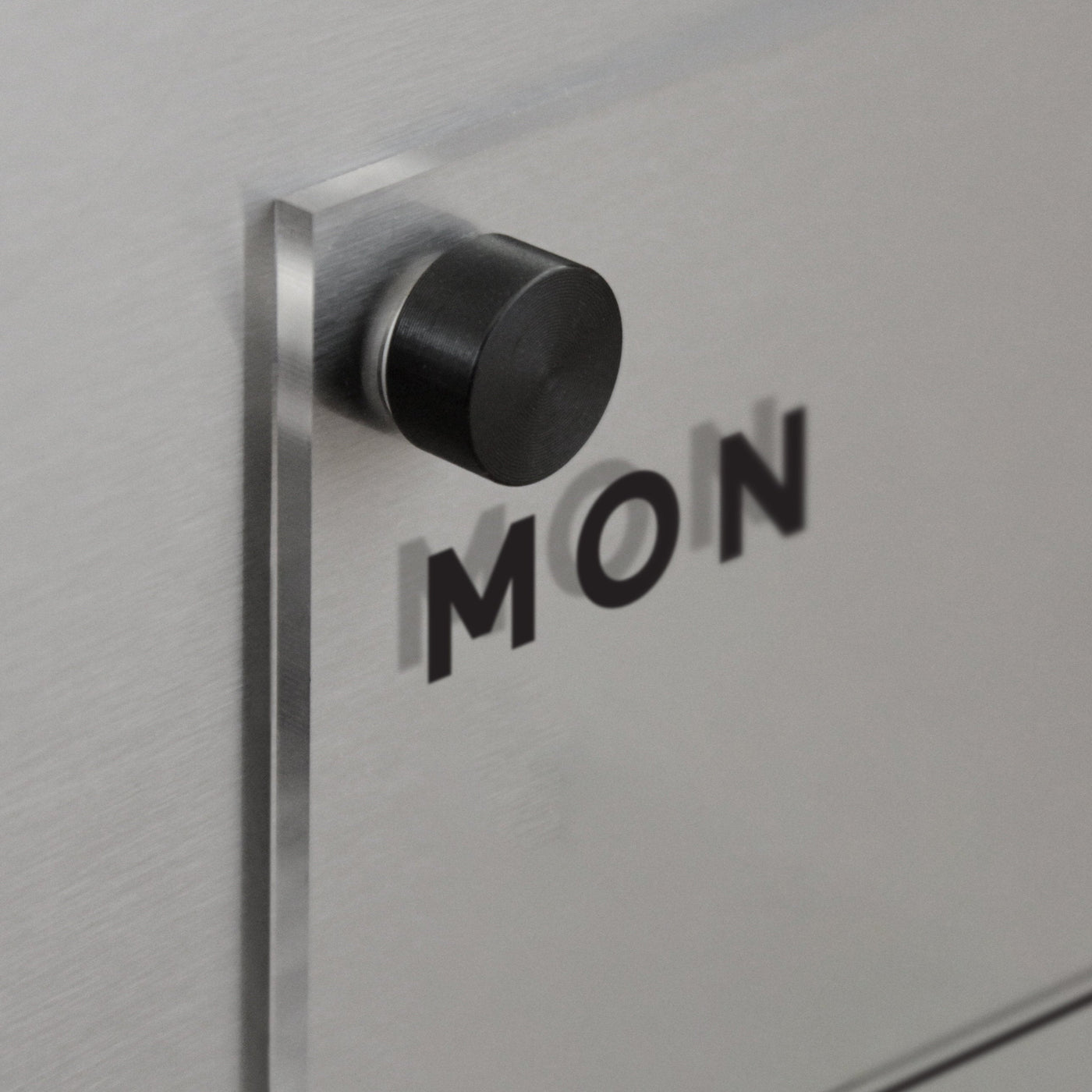 A black circular knob is mounted on a reflective surface resembling the Clear Acrylic Weekly Fridge Calendar by Girl Friday. The word MON is boldly printed with shadow-casting letters, creating a layered effect perfect for wet-erase markers.