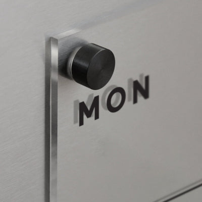 A black circular knob is mounted on a reflective surface resembling the Clear Acrylic Weekly Fridge Calendar by Girl Friday. The word MON is boldly printed with shadow-casting letters, creating a layered effect perfect for wet-erase markers.