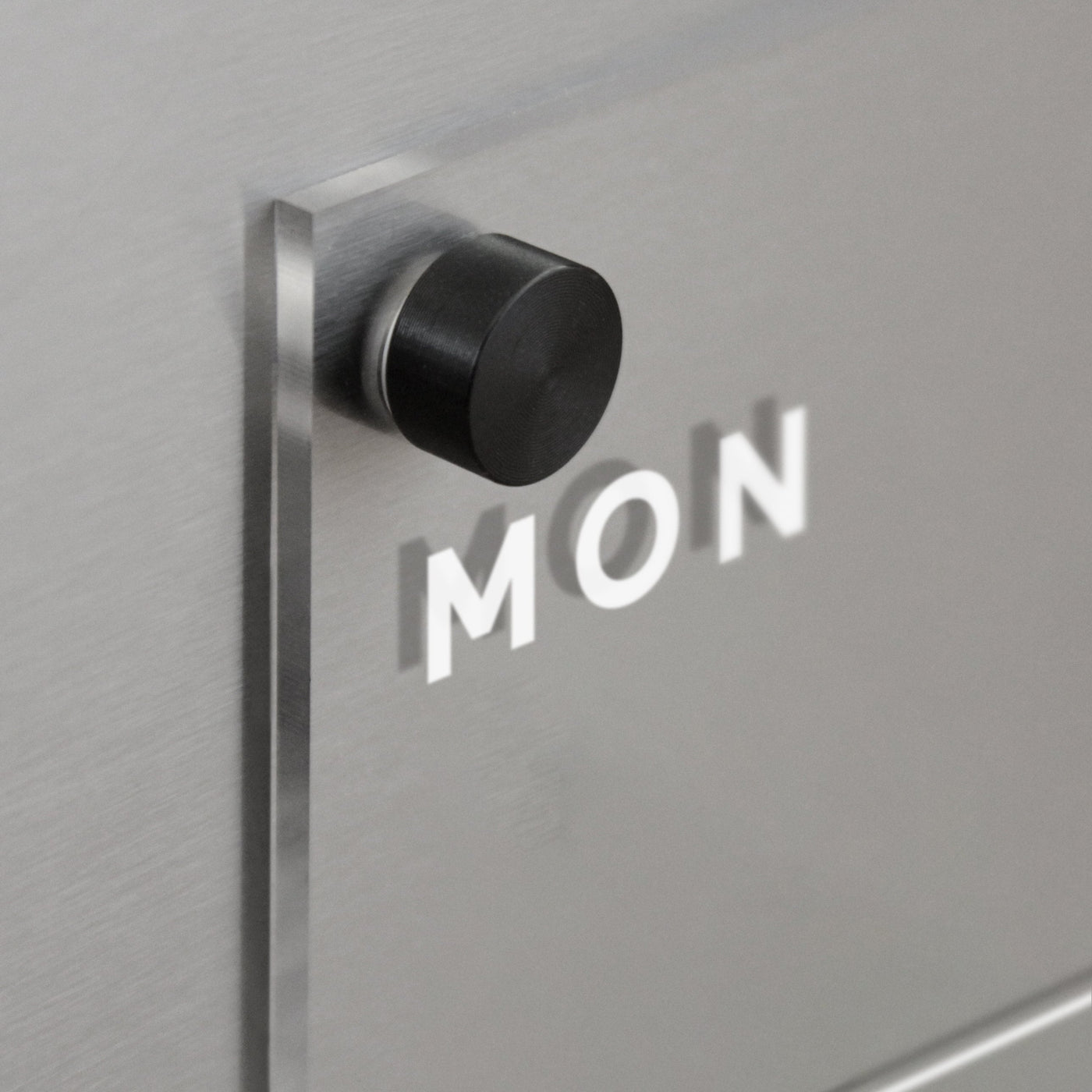 A black round knob on a magnetic surface casts a MON shadow on the sleek, silver metallic finish of the Clear Acrylic Weekly Fridge Calendar by Girl Friday. The shadow is distinct against the smooth background, perfect for notes with wet-erase markers.