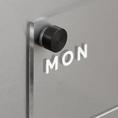 A black circular knob on a brushed metal surface casts a shadow spelling MON. Nearby, Girl Fridays Clear Acrylic Weekly Fridge Calendar (Horizontal, White Text) awaits creativity, ready for wet-erase markers or liquid chalk ink markers to color your week with plans and notes.