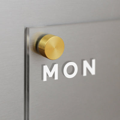 Close-up of Girl Fridays Clear Acrylic Weekly Fridge Calendar with MON in white text, mounted via a round, brass-colored knob on a gray magnetic surface. The design is sleek and modern.