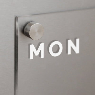 A close-up photo depicts the Girl Friday Clear Acrylic Weekly Fridge Calendar attached to a magnetic surface with a round metal fastener. MON is etched in white on the glass, perfectly suited for use with wet-erase markers.