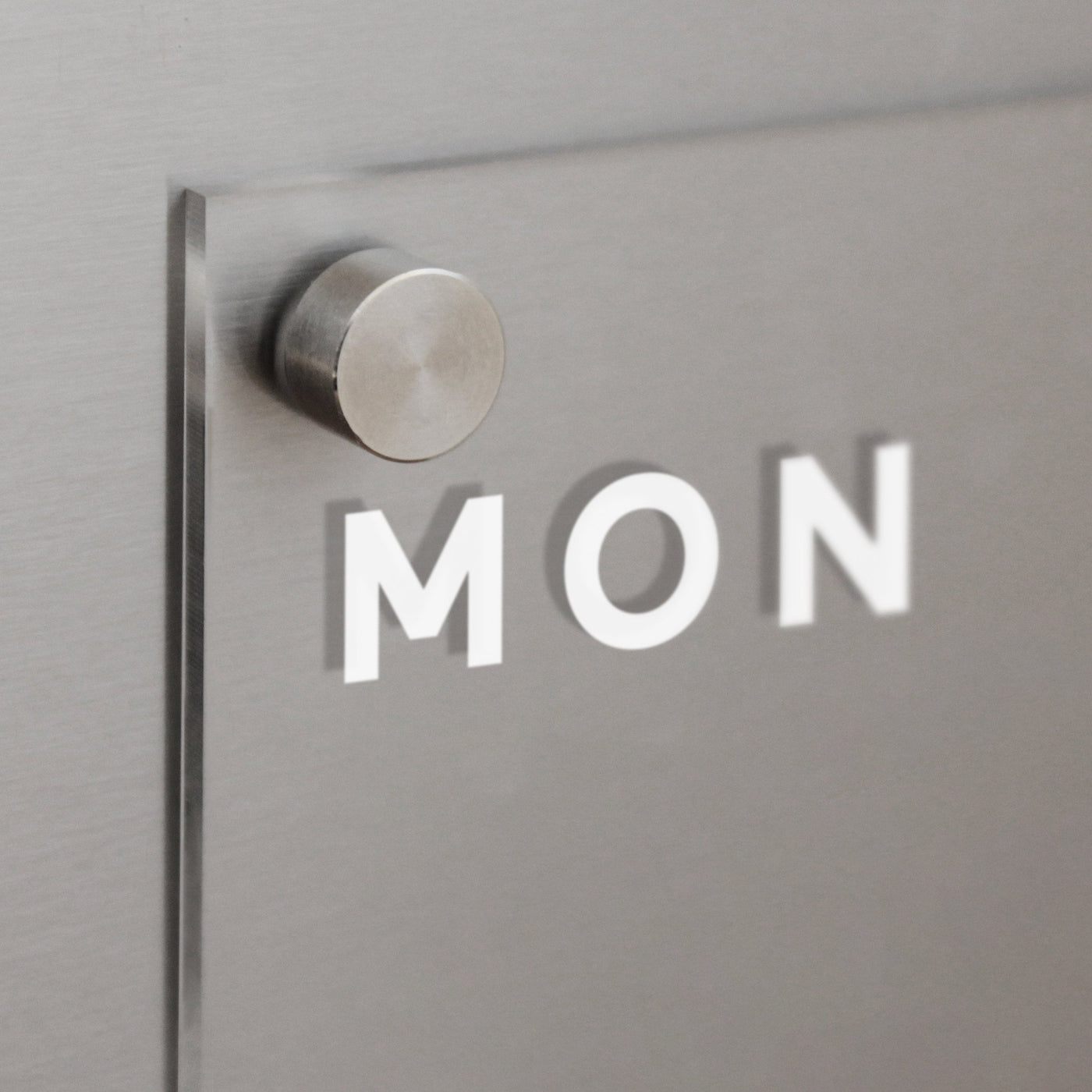 The Clear Acrylic Weekly Fridge Calendar by Girl Friday showcases MON in white on a metallic gray background, with a circular peg for wet-erase markers, offering a minimalist modern design.