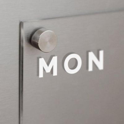 The Clear Acrylic Weekly Fridge Calendar by Girl Friday showcases MON in white on a metallic gray background, with a circular peg for wet-erase markers, offering a minimalist modern design.