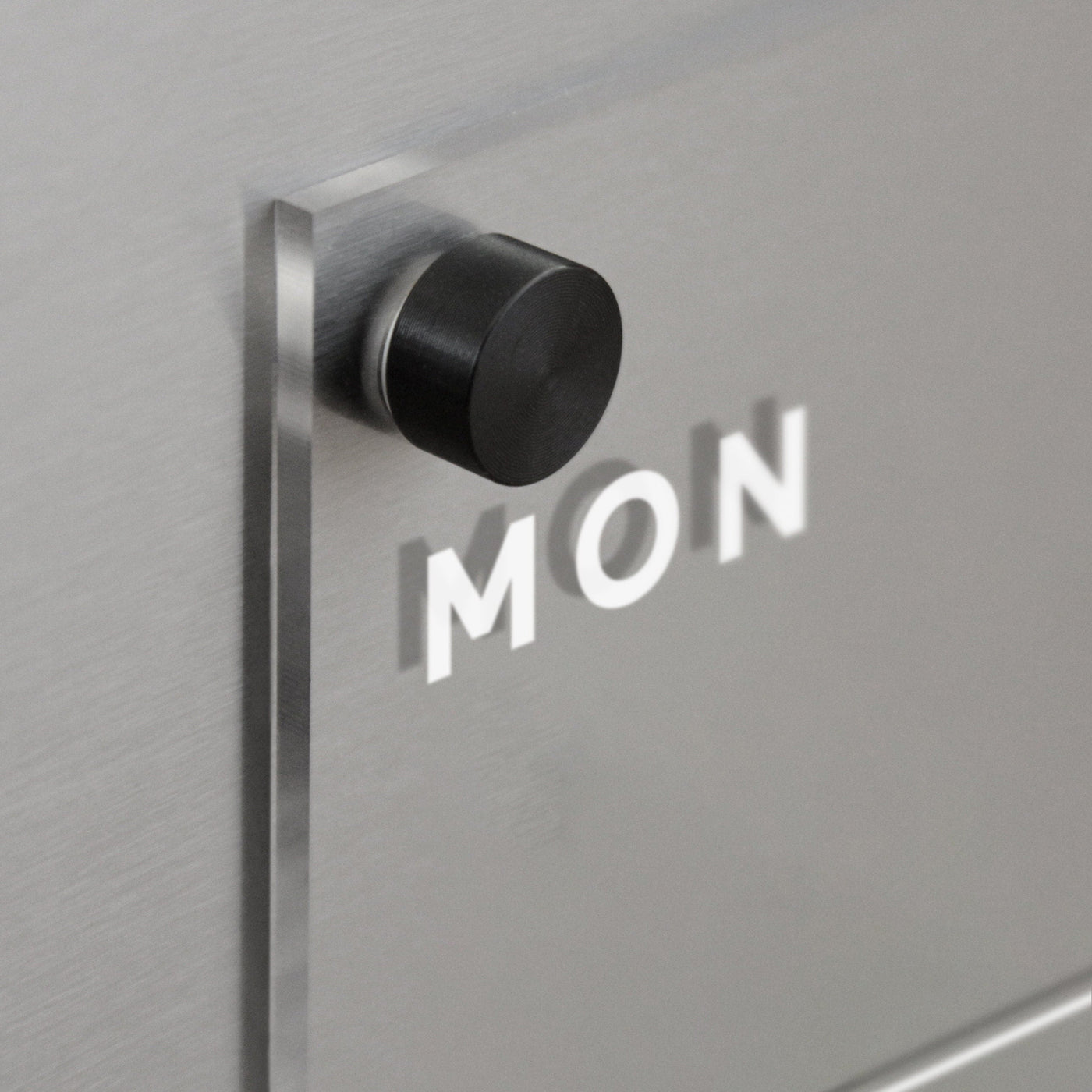 A cylindrical black knob on a magnetic surface casts a shadow spelling MON. The minimalistic design by Girl Friday, ideal for the Acrylic Fridge Calendar weekly, features a brushed metal texture, creating a modern and sleek aesthetic.