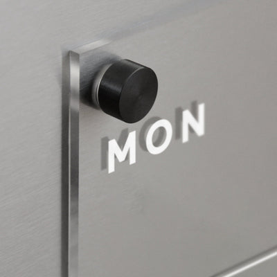 A black knob on a metallic surface casts a shadow spelling MON in white, against a sleek, reflective silver tone. The design is minimalistic and reminiscent of Girl Friday’s Clear Acrylic Fridge Calendar | Week and Notes, ideal for arranging number magnets seamlessly.