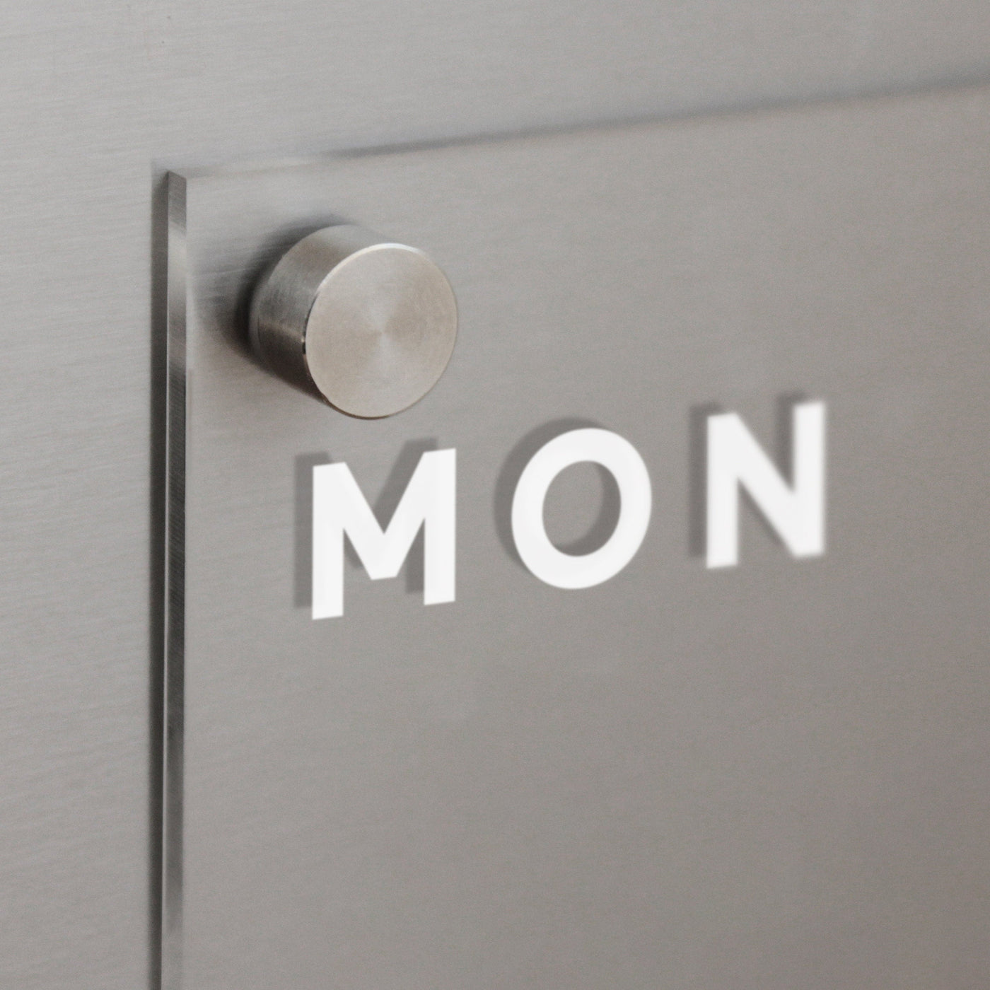 The Girl Friday Clear Acrylic Fridge Calendar | Week and Notes features a metallic wall mount with a frosted glass panel displaying MON in bold white letters. It includes wet-erase markers, providing a brushed metal surface for flexible weekly planning.
