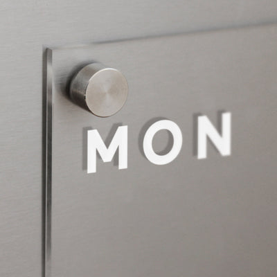 The Girl Friday Clear Acrylic Fridge Calendar | Week and Notes features a metallic wall mount with a frosted glass panel displaying MON in bold white letters. It includes wet-erase markers, providing a brushed metal surface for flexible weekly planning.