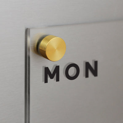 A close-up shows a Girl Friday Clear Acrylic Weekly Fridge Calendar with MON in bold black letters on frosted glass. Its mounted on a silver-gray surface with a gold-tone fixture.