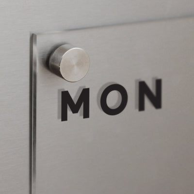 A close-up of the Clear Acrylic Weekly Fridge Calendar by Girl Friday highlights bold black MON letters on a glass panel secured by stainless steel standoff, set against a brushed metal backdrop. Its perfect for wet-erase markers and any magnetic surface, exuding a modern vibe.