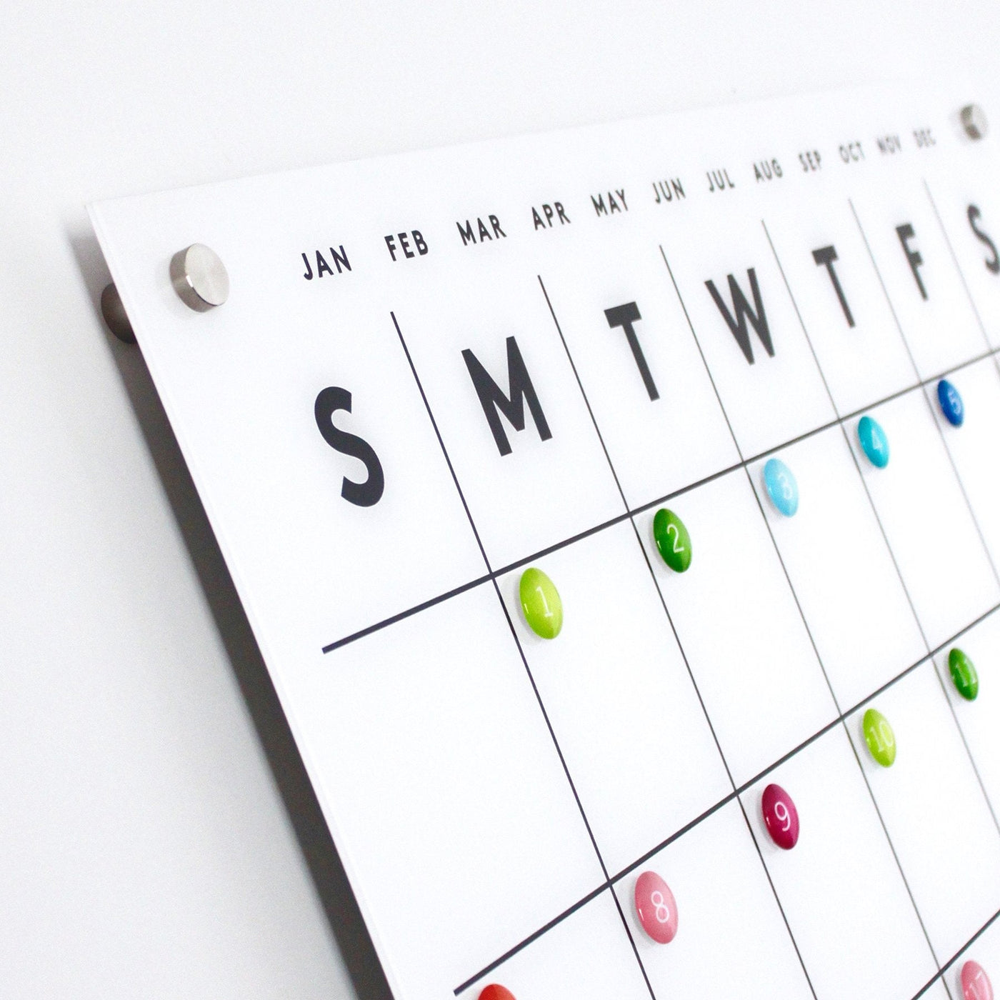 The Magnetic Acrylic Calendar with Side Notes by Girl Friday Shop features a dry erase surface, magnetic capabilities, and colored pins for marking important dates. With days of the week and months clearly displayed, it offers an elegant side angle view.