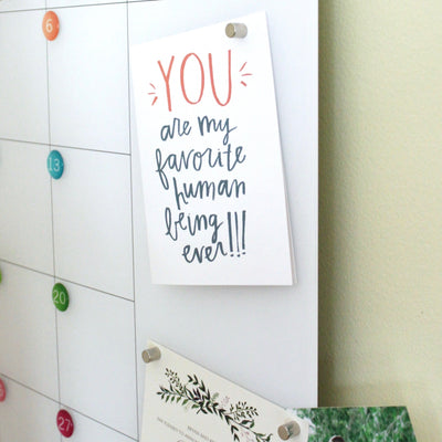 A calendar featuring colorful dots is secured with Girl Fridays Ultra Strong Neodymium Magnets. On the right, a card says YOU are my favorite human being ever!!! in red and black text, while a floral-themed card underneath adds charm to this fridge display.