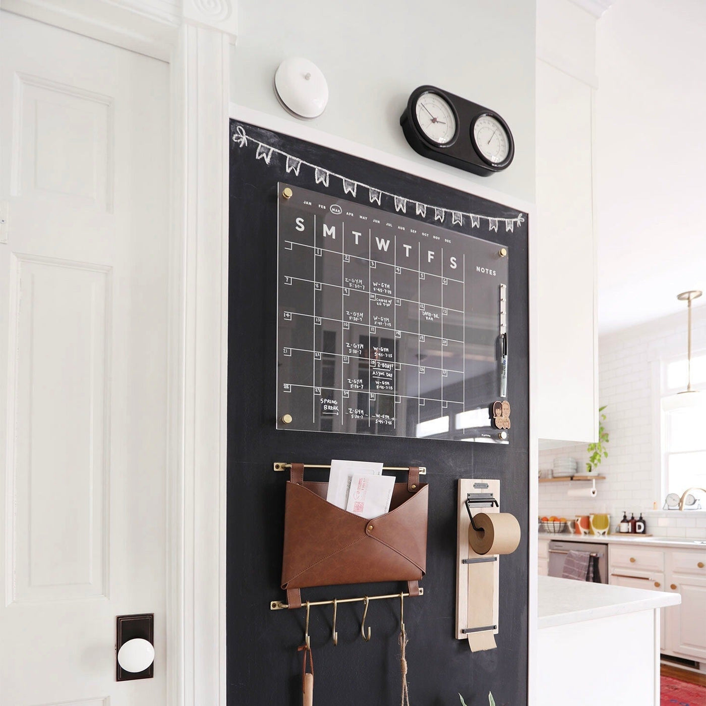 A wall organization setup features the Girl Friday Shops Acrylic Calendar with side notes, displaying white text on a chalkboard background. A brown leather pocket for mail and key hooks complement the design, while a holder with a brown paper roll completes the look against a white door in the kitchen.
