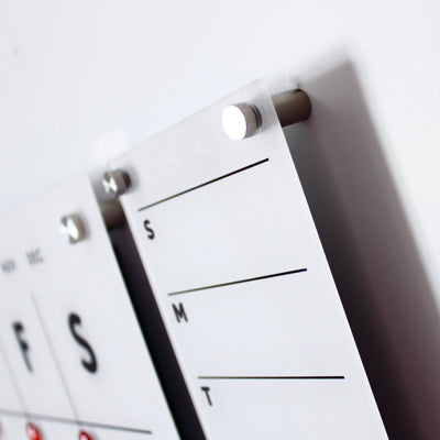 Close-up of a Girl Friday Shop Magnetic Acrylic Menu Board or To Do Board with a modern white wall calendar design featuring black lines and text. Metal pegs hold the clear acrylic panels, and S and M represent days of the week. The minimalist design emphasizes simplicity and functionality.