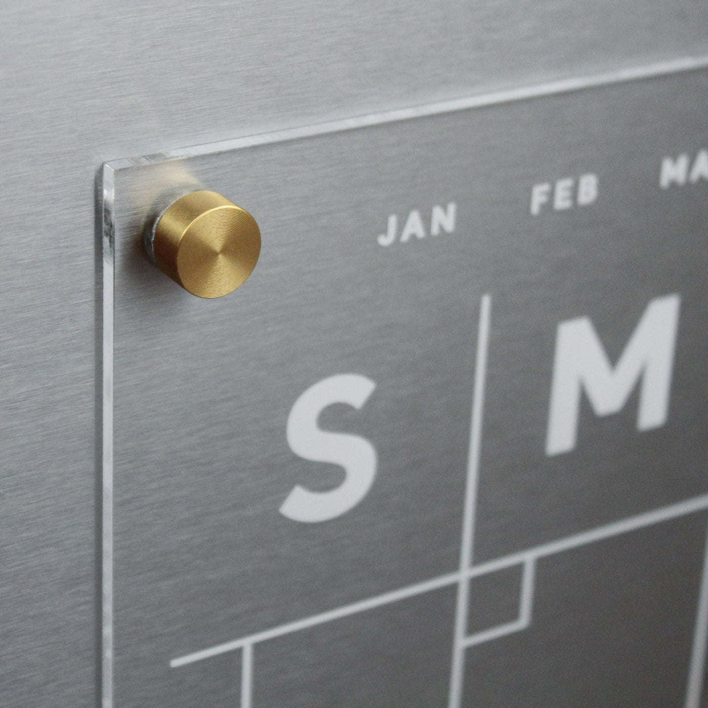 Close-up of the Clear Acrylic Monthly Fridge Calendar by Girl Friday Shop, featuring a sleek metallic design. Days are marked by bold S and M, with visible months like JAN and FEB. A gold cylindrical knob secures the clear panel, ideal for wet-erase markers.