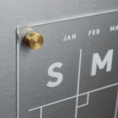 Close-up of the Clear Acrylic Monthly Fridge Calendar by Girl Friday Shop, featuring a sleek metallic design. Days are marked by bold S and M, with visible months like JAN and FEB. A gold cylindrical knob secures the clear panel, ideal for wet-erase markers.