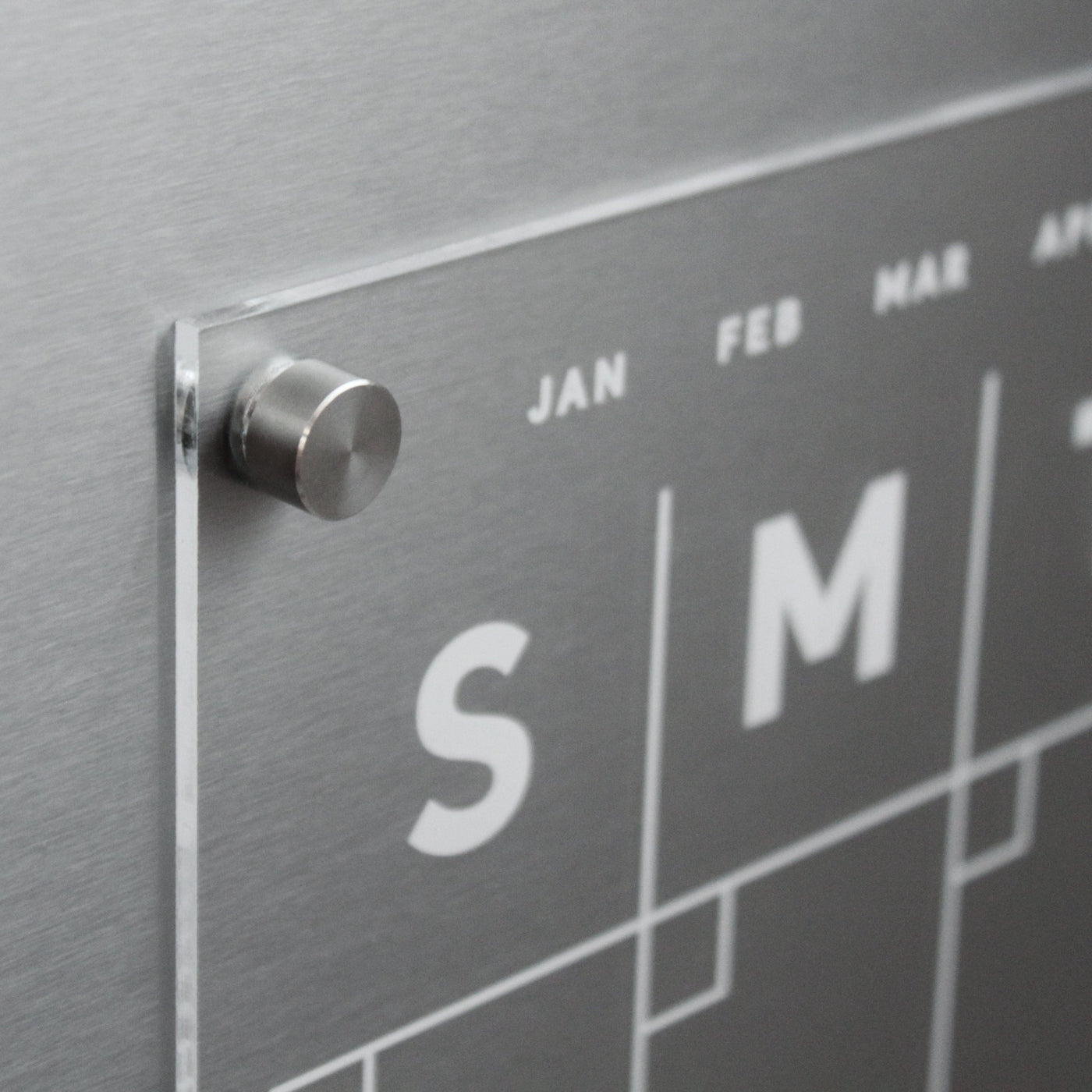 Close-up of Girl Friday Shops metallic, modern Clear Acrylic Monthly Fridge Calendar featuring a glass panel and magnetic surface. Visible are partial week letters S and M plus month abbreviations JAN, FEB, and MAR. Use wet-erase markers on its sleek finish; a metal peg secures it in place.