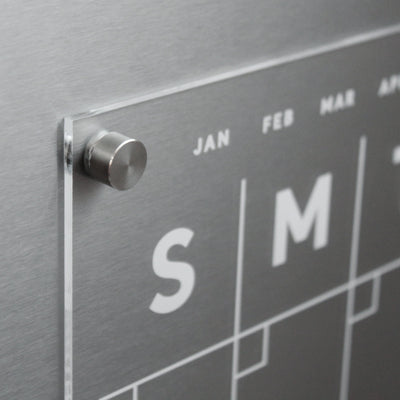 Close-up of Girl Friday Shops metallic, modern Clear Acrylic Monthly Fridge Calendar featuring a glass panel and magnetic surface. Visible are partial week letters S and M plus month abbreviations JAN, FEB, and MAR. Use wet-erase markers on its sleek finish; a metal peg secures it in place.